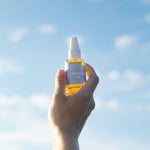 clerstory glow oil - radiant body oil, sunflower and citrus, vegan plantbased cruelty-free product being held by a hand in front of a blue sky on a sunny day; golden oil in a clear bottle with blue label