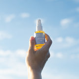 clerstory glow oil - radiant body oil, sunflower and citrus, vegan plantbased cruelty-free product being held by a hand in front of a blue sky on a sunny day; golden oil in a clear bottle with blue label